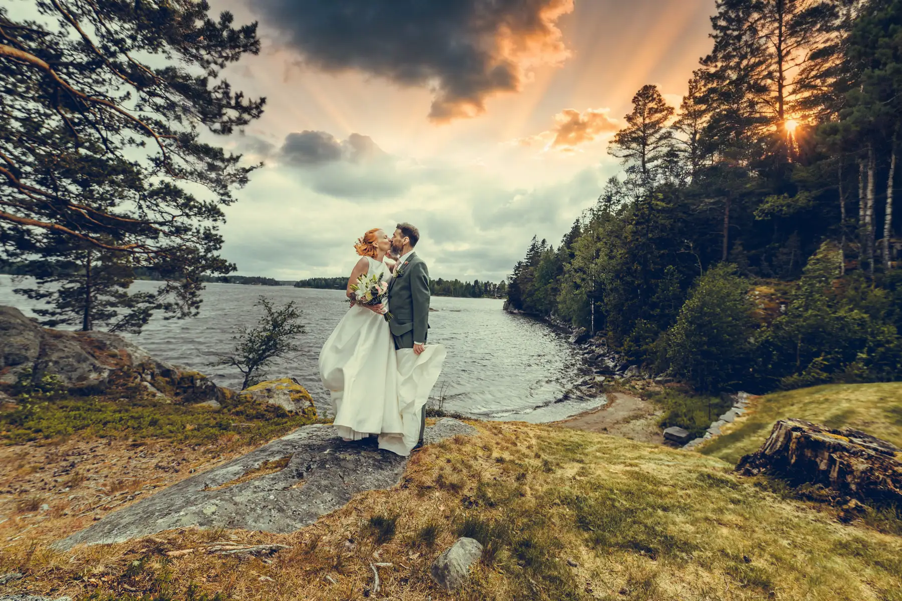 Bryllupsfotograf Oslo – brudepar ved vann omgitt av skog og solstråler gjennom mørke skyer
