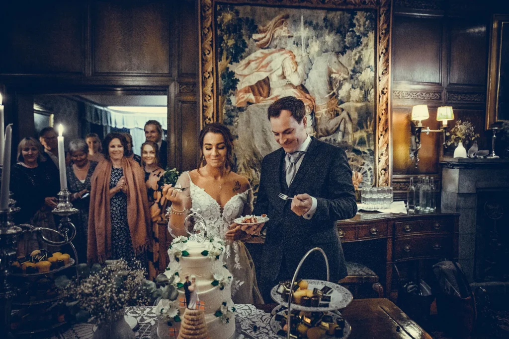 Bryllupsfotograf Oslo – brudepar ved bryllupskake omgitt av gjester i elegant sal, øyeblikksbilde med smil og feststemning