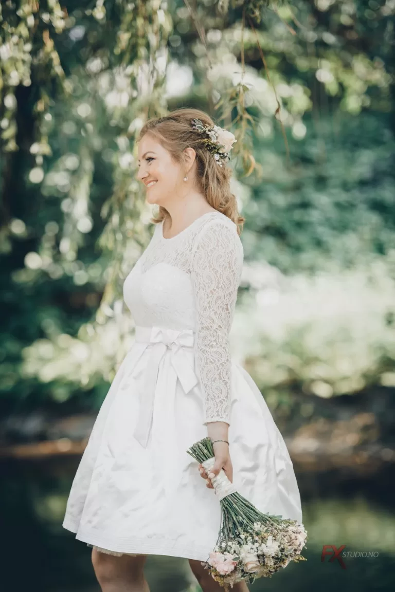Bryllupsfotograf – detaljert bilde av brud med kort kjole og blomsterbukett, fotografert på Nedre Foss Gård i dokumentarisk stil ved vann og grønn vegetasjon