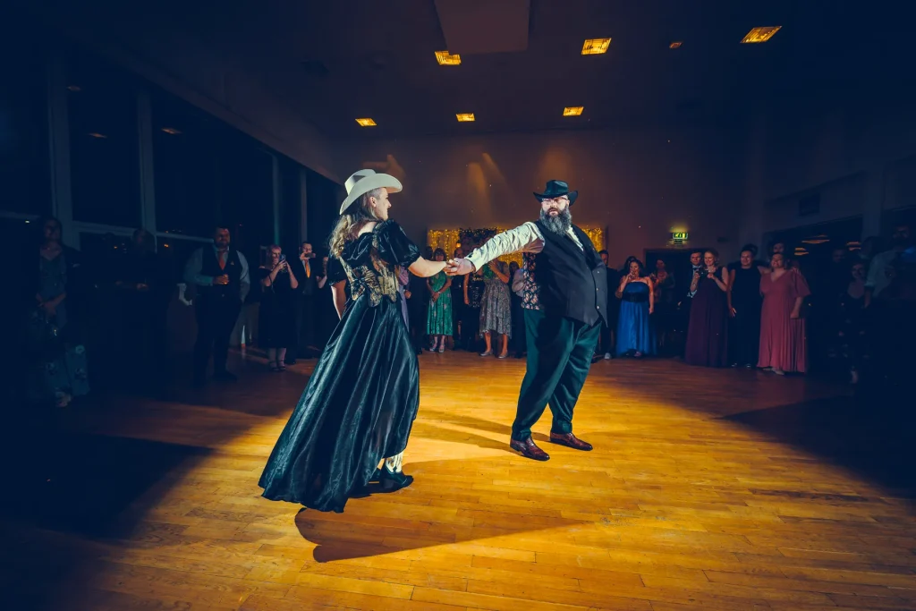 Dansende par i spotlight under bryllupsfest på Sørskogbygda Samfunnshus – kvinnen i grønn kjole med blomsterdetaljer og cowboyhatt, mannen i vest og hatt, omgitt av festkledde gjester.